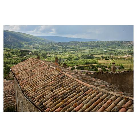 Wide angle image looking over the tiled rooftops of a village in Provence, France. Green fields and vineyards are visible in the distance where they meet a mountain range. The sky is light blue with clouds.