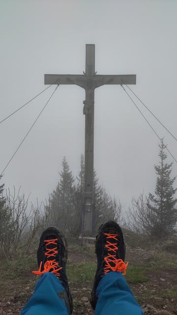 Gipfelkreuz des Immenstädter Horn umgeben von grauem Nebel.
Im Vordergrund ein paar ausgestreckte Beine mit bläulichen Hosen und schwarzen Wanderschuhen mit orange farbenen Schnürsenkeln.

Summit cross of the Immenstädter Horn surrounded by grayish fog. Stretched out feet with bluish pants and black hiking boots with orange laces are in the foreground.