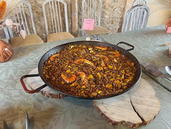 A paella style wide and flat pan on a table. One can see prawns, clams, and other seafood mixed in amongst brown noodles.