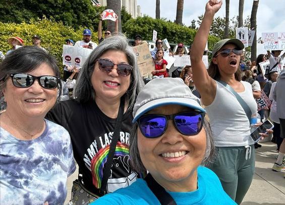 Several women smile in a selfie, surrounded by sign-waving, fist-raising protestors