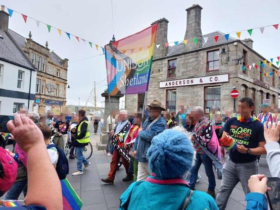 A colourful parade marching in front of grey old buildings. In the centre two people carrying a huge Pride banner with the text 'Unison Shetland'