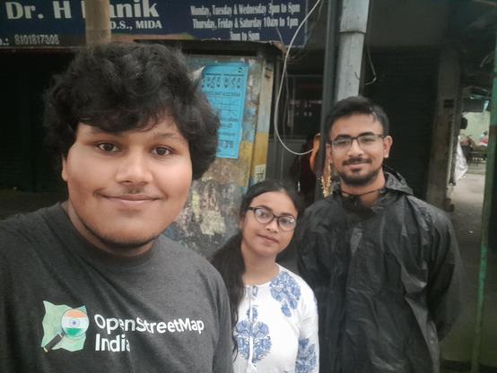 2 men and a woman standing next to a dentist's office in Kadamtala, facing the camera. The leftmost person in the picture is wearing a grey OpenStreetMap India T-shirt.