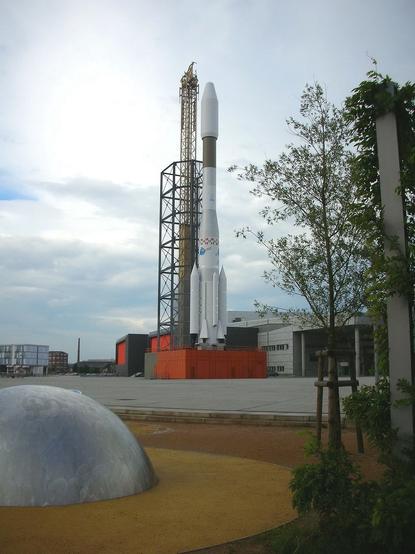A large rocket is depicted on a launch pad, accompanied by a tall scaffolding structure. In the foreground, there is a metallic dome on a gravel surface, with trees and modern buildings visible in the background under a cloudy sky.