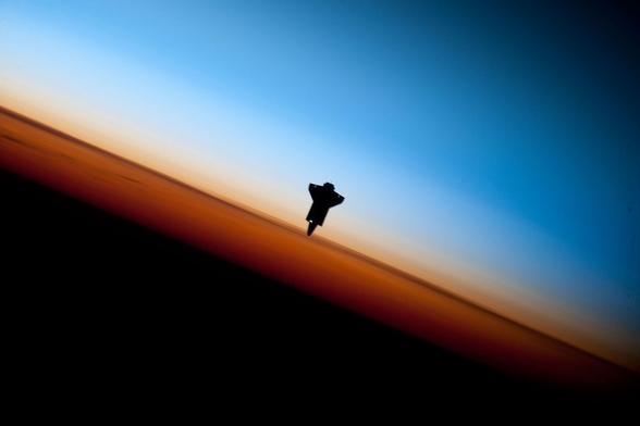 The image shows a space shuttle silhouetted against a gradient sky transitioning from blue to orange, likely during dawn or dusk, with a dark foreground.