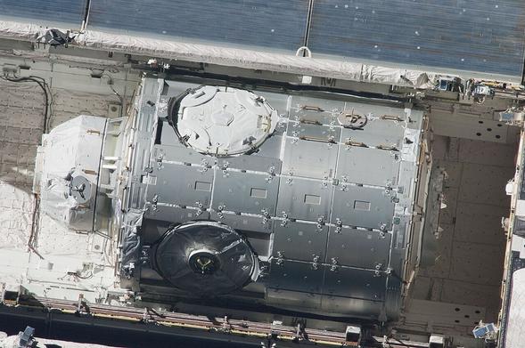 An aerial view of a spacecraft module with a shiny metallic surface, mounted within a larger structure. The image showcases intricate details, including circular openings and a panel design, set against the backdrop of the spacecraft's interior.