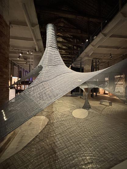 Large architectural textile structure suspended inside a historic hall. The translucent woven fabric forms flowing, organic shapes: a cone rising towards the ceiling, a column, and stretched surfaces anchored to walls and floor. The mesh casts intricate shadows on the ground, creating patterns that echo its geometry. The material appears both delicate and resilient, inviting reflection on tension, balance, and temporality.