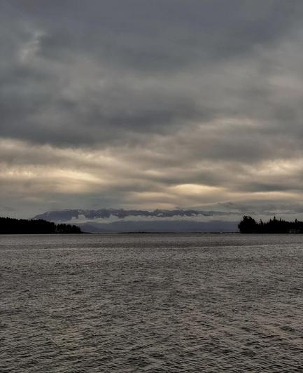A landscape photograph captures a moody seascape under a dramatic sky. The dark, rippling water fills the foreground, leading to a distant shoreline with silhouetted trees. Beyond the shoreline, a low mountain range is partially obscured by clouds. Above, a heavy, layered sky features varying shades of gray, with hints of lighter areas peeking through the cloud cover.