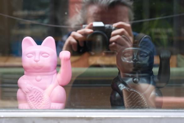 Hinter einem Schaufenster steht links eine rosafarbene winkende Glückskatze und rechts eine schwarze, weniger auffällige, als wäre sie ein Echo der ersteren; im Glas sieht man ein verschwommenes Spiegelbild der Person, die das Foto macht, ein Echo von mir selbst. / Behind a shop window are a pink fortune cat, waving on the left, and, on the right, there is a black one, less noticeable, as if it were an echo of the former; in the glass you can see a blurry reflection of the person taking the picture, an echo of myself.
