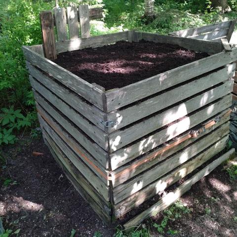 Puinen kompostikehikko täynnä multaa. Kehikon ympärillä kuormaliina ja takana puolikkaaksi lahonnut kehikonseinä pystyssä.