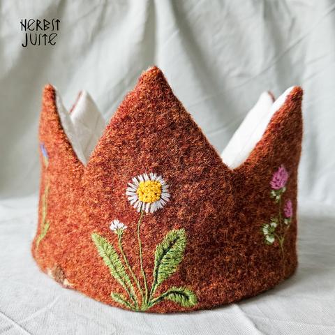 Eine Krone aus rostrotem Wollwalk (außen) und weißem Leinen (innen). Sie ist realistisch bestickt. Aus dieser Perspektive sieht man sie von vorn, gestickt ist ein Gänseblümchen.