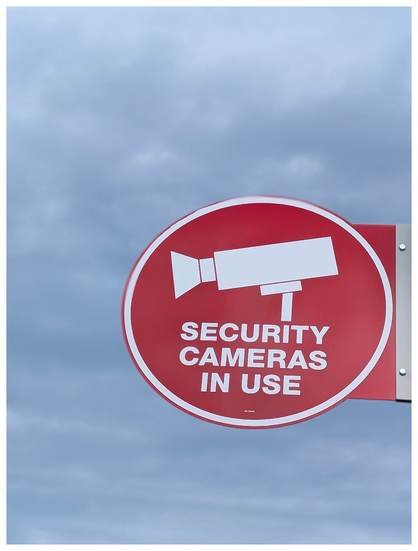 A red circular sign with a white graphic of a security camera and bold white text reading “SECURITY CAMERAS IN USE” is displayed against a cloudy blue sky.