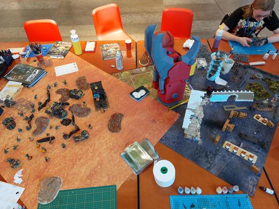Top view of the table with desert terrain mat with desert scenery and corrugated iron Buildings, barriers and orcs versus kroots shooting at one another, and on the other side a ruined town mat with working progress fantasy buildings. In the centre is an obscenely large 3d printed power claw everyone wanted to be on a photo with