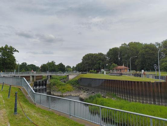Die Schleuse Dömitz bzw. der Bereich mit der Fischtreppe davor. Rostige Spundwände vor teilweise verlandeten Gewässerlauf der Löcknitz. Ringsum Laubbäume. Neben der Fischtreppe ist noch ein weiteres Sperrwerk, wo wenig Wasser durchläuft. Im Hintergrund wehen die Deutschland- und eine Mecklenburgische Fahne.