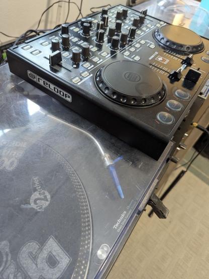 The image shows a DJ equipment setup. In the foreground, there is a transparent turntable with a blue tone arm. On the turntable, a 1210, there is a logo that reads "Technics." Behind the turntable, there is a DJ controller labeled "RELOOP." It is a Reloop Master Edition 2, still slightly dirty, but has been cleaned recently.
