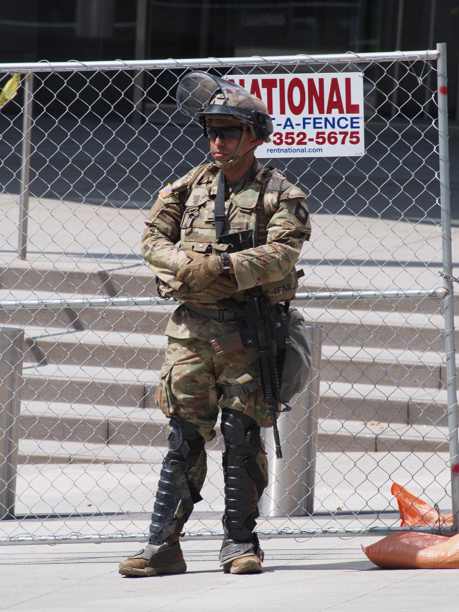Soldier standing in front of a rent-a-fence. He is armed with a rifle, has black shin protection over a camouflage uniform. His helmet has a shield which is tipped back. He is wearing dark sunglasses and has his arms crossed.