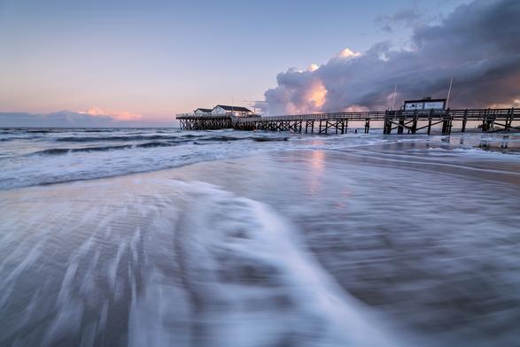 Zu sehen ist eine Aufnahme in Langzeitbelichtung von der Nordseeküste, bei der im Vordergrund das Wasser durch den Wind gepeitscht wird. Im Mittelgrund das Meer sowie ein Restaurant auf Bunen und im Hintergrund bewölkter Himmel.