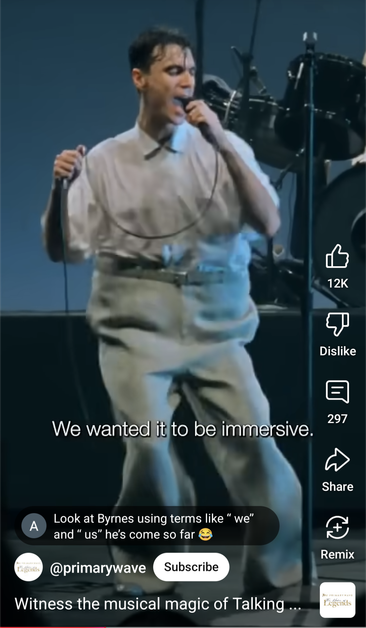 Video screenshot. A much younger David Byrne sings into a mic on stage, in an oddly bulky outfit, weirdly wiggling his hips.