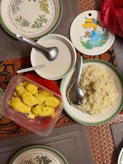 Top down photo of a dining table with large plate of Pulut (sticky rice), a bowl of Santan (Coconut milk), and a plastic tub full of yellow, ‘fatty’ Durian.