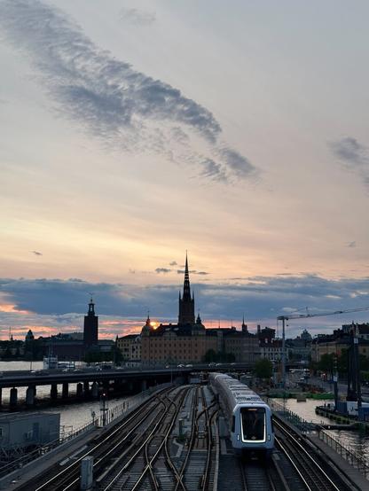 A view over a span of six railway lines receding into a tunnel in the distance, above which is an old European city. The sun is setting in the distance. A modern metro train, its sides glossy white like a Star Wars Stormtrooper suit, its front illuminated with a square U of white LED strips, drives forth on the second track from the right.