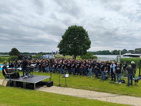 Eine Wiese aus einem anderen Blickwinkel in einem Beachclub. Auf der Wiese stehen ca. 170 Chorsängerinnen und Sänger aus den verschiedensten Rockchören60+. Geleitet wird das Event von Dieter Falk
