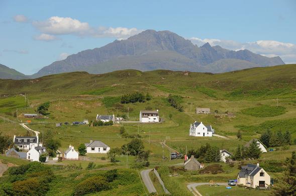 Tarskavaig on the Isle of Skye. The image shows a scatter of white houses in the foreground leading back to an open area higher up a hill and then a horizon. Beyond, in the distance is a jagged mountainous ridge. The scene is in sunlight.