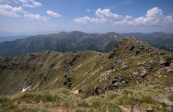 Im Vordergrund Gras, dahinter ein Bergkam nach links führend, ein Tal und weiter hinten weitere Berge. Es gibt einige Wolken, der Himmel ist blau.