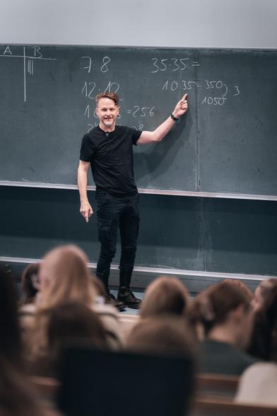 Christian vor der Tafel mit einigen Rechnungen darauf im Hörsaal