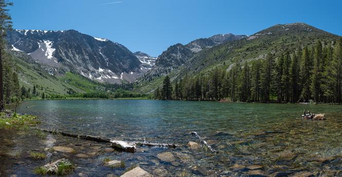 Parker Lake, near June Lake, Caifornia, looking across the lake to mountains still with some patches of snow in the more sheltered areas.  The lake is very clear, bolders seen through the close water, and some logs.  Trees on the shore to the left of frame, and a U shaped valley in the far distance, meadows and trees stretching up the valley.