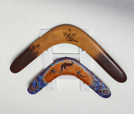 Eine der weißen Halterungen hängt an der weißen Wand.
Sie trägt zwei australische Boomerang aus Holz. Verziert und bemalt von Aborigines-Künstlern.
Es sind auch Kängurus auf den Flugwinkeln zu sehen..