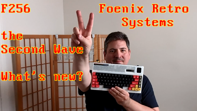 Picture of me holding up a wedge computer F256K2 in one hand, and holding up 2 fingers witht the other hand.  Red letters in yellow outline spell out: "F256    Foenix Retro Systems, the Second Wave. What's new?"