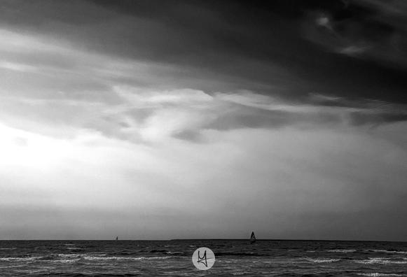 Das Bild zeigt eine ruhige Meereslandschaft in Schwarz-Weiß. Im Vordergrund sieht man sanfte Wellen, die an den Strand rollen. Am Horizont erkennt man zwei Windsurfer, die sich auf dem Wasser befinden. Der Himmel ist von dramatischen, teils dunklen Wolken und helleren Wolkenformationen durchzogen, was dem Bild eine stimmungsvolle, fast melancholische Atmosphäre verleiht. Insgesamt wirkt die Szene ruhig und weitläufig, mit einem starken Kontrast zwischen Himmel und Meer.