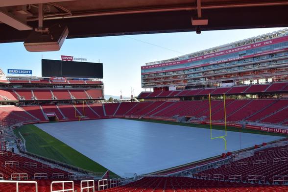 View from a stadium interior showing red seats, a central green field partially shaded, and a large digital scoreboard at one end. The stadium has multiple levels with exposed walkways and seating areas. Columns of iconic banners noting 'Super Bowl Champions' hang prominently. The field is marked in one corner with a yellow goal post. Bright sunlight illuminates the scene, casting shadows across the seats.