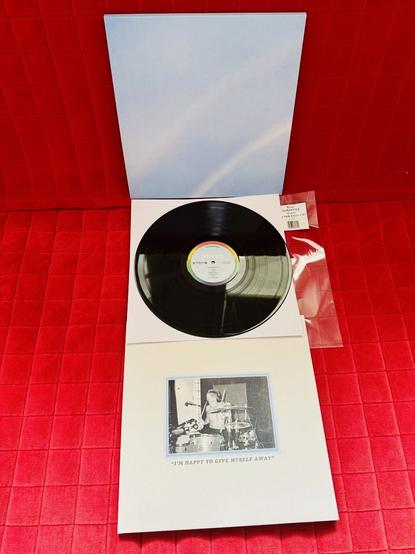 The image shows a vinyl record edition of "Never Enough" by the band Turnstile. The record is placed on a bright red textured surface. The vinyl is black with a white center label that features the album title "Never Enough" and some small text around the edge.

The open gatefold cover reveals two parts:

    The top panel is a soft, light pastel blue with a faint rainbow arc — a minimalist and dreamy design.

    The bottom panel shows a black-and-white photo of a shirtless drummer playing passionately, framed in pale blue. Below the image is a quote in capital letters:
    “I’M HAPPY TO GIVE MYSELF AWAY”

To the right of the record is a clear plastic sleeve with a barcode and a white sticker label reading:
"TURNSTILE - NEVER ENOUGH"