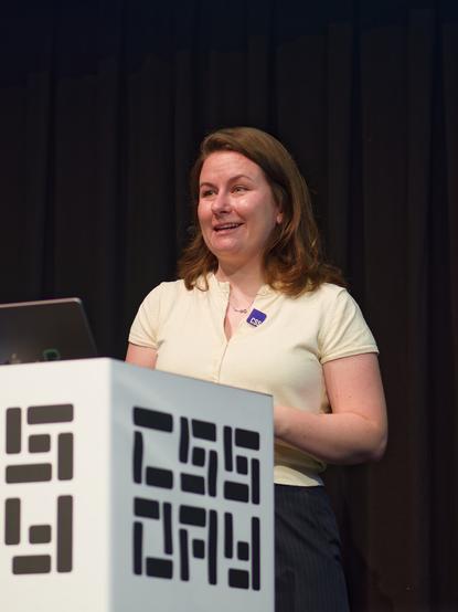 A photo of Cyd, standing near a podium with a “CSS Day” logo on it, giving a talk while smiling and looking at the audience. There is a MacBook with a few logos on its lid on the podium. Cyd is wearing a pale yellow shirt and a CSS logo pin on it.