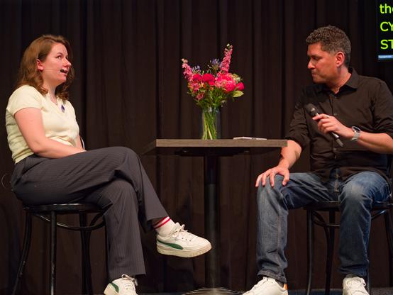 A photo of Cyd and Stephen sitting on chairs on the scene. Stephen is looking at the phone with the questions lying on the table near a vase with colorful flowers, and Cyd is looking up and answering some question.