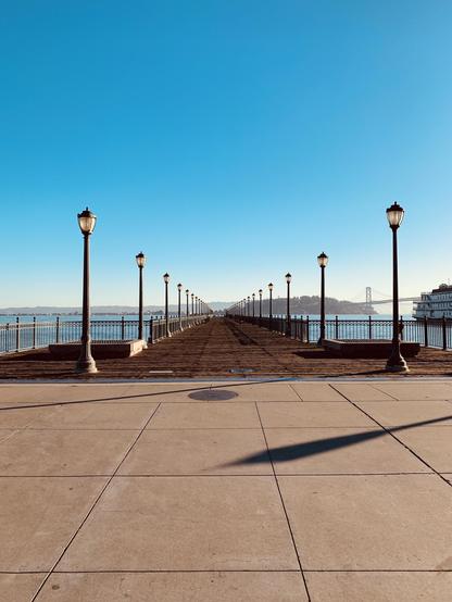 Long pier paved with beige tiles and lined with old-fashioned lamp posts, leading towards a body of water under a clear blue sky. In the distance, a partial view of a bridge is visible.