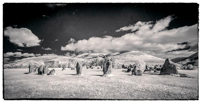 A toned black and white infrared photo showing a circle of various sized upright boulders in an area of short grass, lit by low angle sunlight, with clouds and a blue sky.