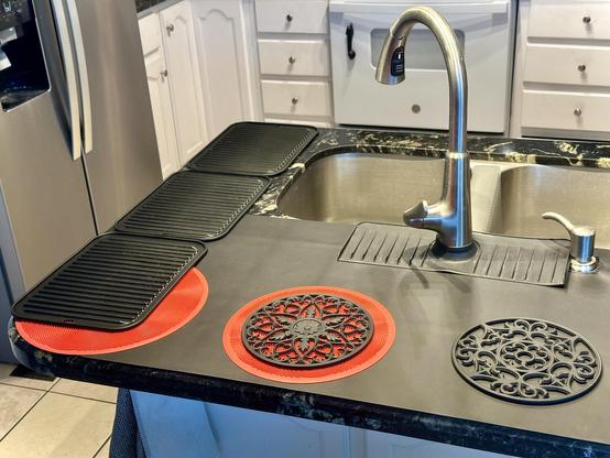 After photo shows peninsula/island sink area has silicone mats to protect the black granite counters from water or hot pans. All black thin black mat covers most of the large counter area behind the faucet. 3 ribbed heavy silicone trivets line the left counter edge. 2 red silicone mats are at the corner facing me, placed under black silicone trivets. 2 round decorative black silicone trivets towards center and bottom right of photo. A little of the kitchen seen on 3 sides of the sink area:fridge left, white stove and drawers top, wood white cabinet under sink area counter.