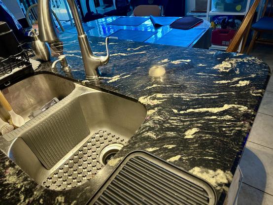 Before photo. Ooh black granite counters show in this photo of the peninsula/island sink area. 2 deep stainless sink tubs unit with single post tall faucet and lever. Dish soap dispensers to top left of faucet. Dining area shows beyond sink and table. It looks nice but water constantly splashes counter in a wide area.