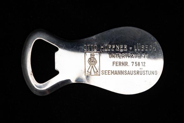 This is a photo of a vintage metal combination bottle opener and shoe horn stamped with the name and details of a company: "Otto Höpfner – Lübeck, Untertrave 70, Fernr. 75812, Seemannsausrüstung" (which translates to "seamen's equipment"). The opener also features a small logo of a stylized figure with the initials "OH" on its chest.