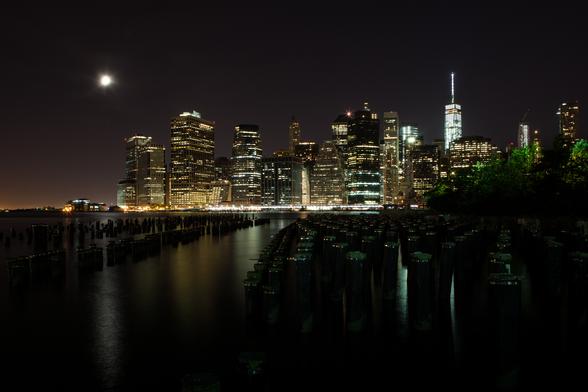 A nighttime skyline of a city with illuminated buildings, a bright moon in the sky, and old pier pilings in the water in the foreground.