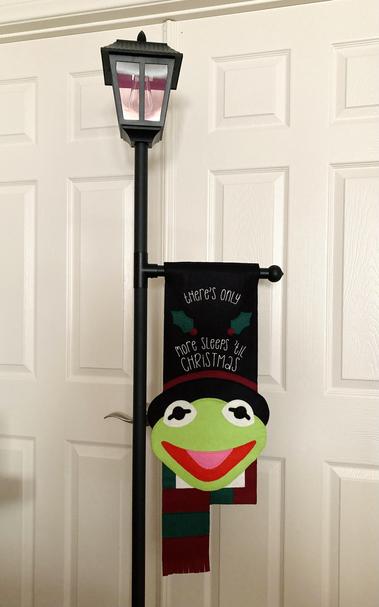 A Victorian looking lamppost with a cross bar that holds a long felt Advent calendar shaped like Kermit in a top hat from A Muppet Christmas Carol. His top hat is exaggeratedly tall and has been machine embroidered in white with the phrase "there's only ... more sleeps 'til Christmas" with space for a felt number to be attached between two blanket stitched holly leaves