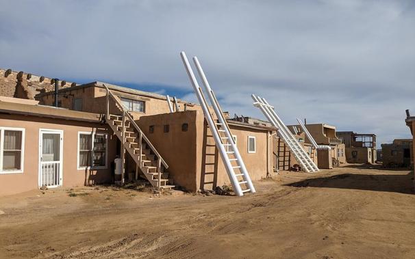 A line of 2-story adobe pueblo houses, all painted the usual orange-brown, with white window and door sills, on a wide dirt lane. Second from the left is a pueblo that has a ladder propped against it, with the sides made of wide round poles, painted bright white and contrasting against the orange-brown of the buildings. The ladder rungs go up to the roof of the bottom story then stop, but the sides of the latter continue much higher. The next house down has a double (side by side) version of the same ladder, and you can see a few other ladder tops protruding from other houses. The nearest house to the camera (far left of the photo) has an unpainted wooden staircase leading up to the roof instead of a ladder.