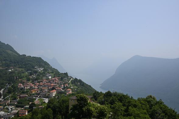 Aussicht auf das Bergdorf Brè vom Monte Brè aus. Im Vordergrund ist ein Dorf mit Steinhäusern mit roten Dächern an einem seichten, bewaldeten Berghang. Im Hintergrund ist eine Verblaute Landschaft mit Bergen an einem Bergsee. Die Verblauung im Hintergrund ist so stark, dass der See und die Berge weiter hinten in den Himmel übergehen.