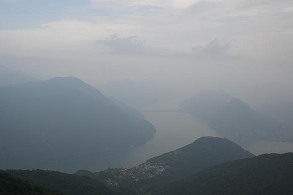 Aussicht auf das Bergdorf Brè vom Monte Boglia aus. Eine Berglandschaft in Blautönen mit starkem Verblauungseffekt. Im Vordergrund ist ein bewaldeter Berghang. In eine Mulde ist ein Bergdorf gekuschelt. Dahinter ist ein verzweigter See, der von Bergen umrundet ist. An einer Stelle im Hintergrund geht eine Landbrücke über den See. Die Verblauung im Hintergrund ist so stark, dass der See und die Berge weiter hinten in den Himmel übergehen. Der Himmel hat verschiedenen Hellblautöne und es sind ein paar Wolken erkennbar.