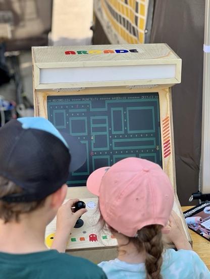 Bild zeigt zwei Kinder mit Mütze vor dem Arcade Automat, die ein Retrogame zocken.