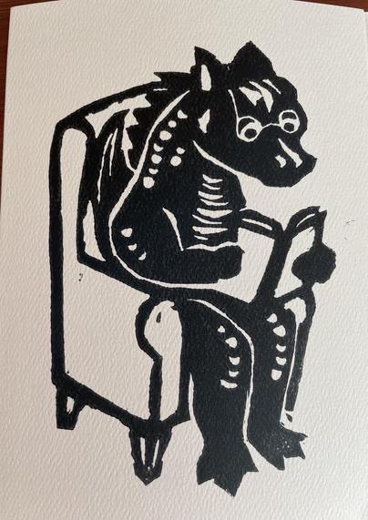 A linocut image on a card of a dragon sitting and reading a book