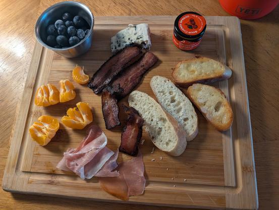 A cutting board, because I am too lazy to do dishes, filled with a cup of blueberries,a mandarin orange,a couple slices of prosciutto, a couple slices of mango habanero bacon, a chunk of Roquefort, some sour cherry spread, and some good french bread, some toasted and some not.