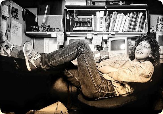 Fotografia dos anos 80 de Susan Kare, uma jovem de cabelo encaracolado pelos ombros, de sweatshirt e calças de ganga, usa peúgas e calça ténis. Está de braços cruzados, reclinada na cadeira de trabalho e pés apoiados na sua mesa com uma perna esticada e a outra semi-dobrada. Ri-se de algo que lhe é dito numa conversa com intervenientes que não foram enquadrados na foto.
Atrás tem estantes com livros de programação, por cima deles há jogos de tabuleiro; à esquerda um pequeno robot de uma série de animação exposto juntamente com óculos antigos, uma garrafa de champanhe.
Na mesa de trabalho em L tem 3 Macs espaçados, por cima do Mac da direita e que está atrás da sua cabeça, o que parece ser uma caixa de legos.
Nas paredes do cubículo: fotografias, anotações, recados, tabelas...
No canto inferior esquerdo, aparece parcialmente uma impressora a 'cuspir' papel contínuo perfurado nas laterais.
