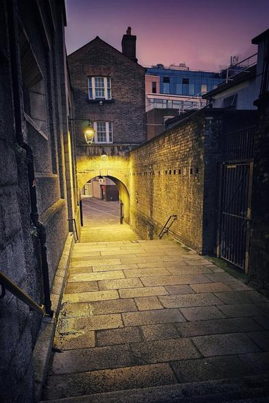 A narrow, historic alleyway in Dublin at dusk, illuminated by warm yellow light from a streetlamp under an archway. The stone steps lead down through the arch to a dimly lit street beyond. On the right, a brick wall is brightly lit, and on the left, an old stone building with a small window is visible. In the background, a modern building with blue windows can be seen against a purple and orange sky.